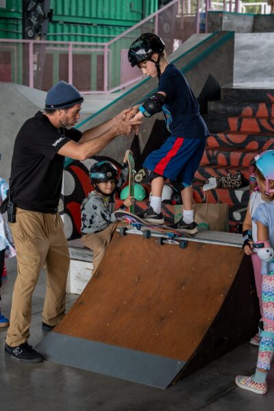 Instructor helps a young boy balance on a skateboard ramp while others wait.