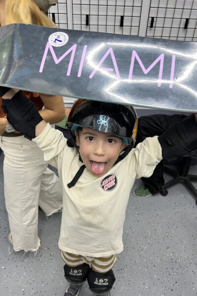 Excited young skateboarder holding a shiny deck labeled “MIAMI” over head.