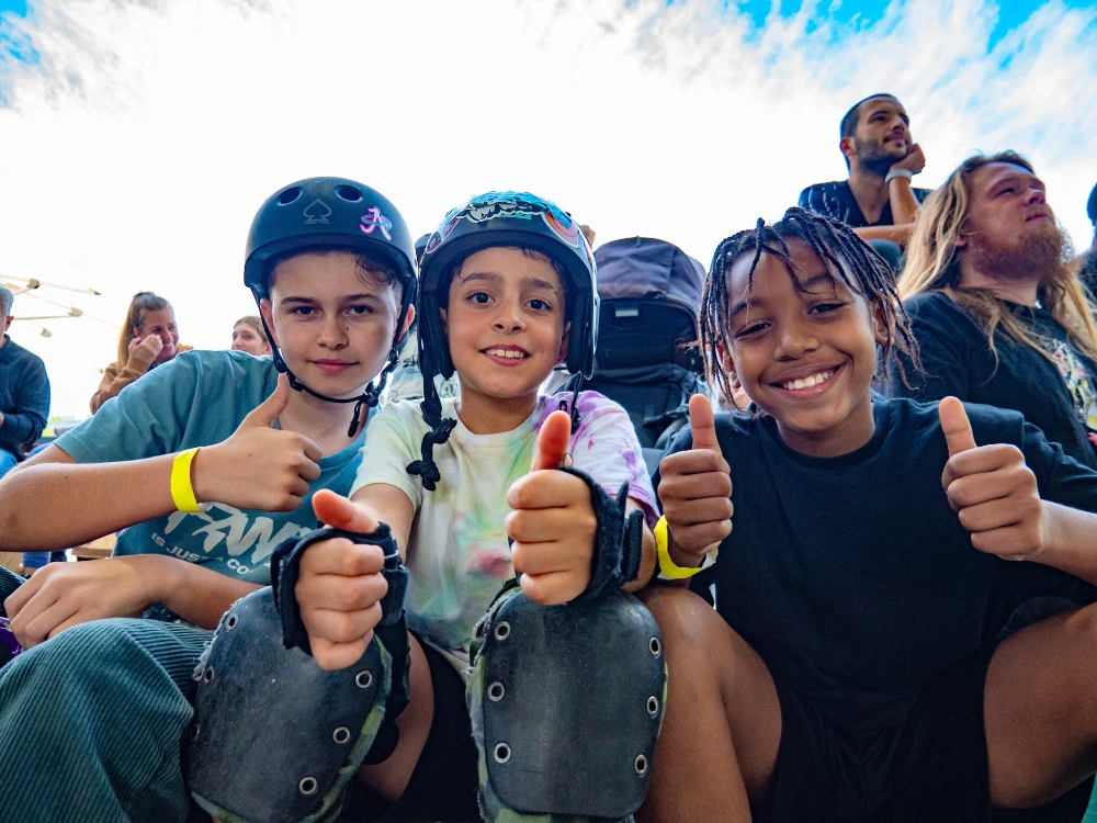 Three happy kids in skate gear give thumbs up while sitting together.