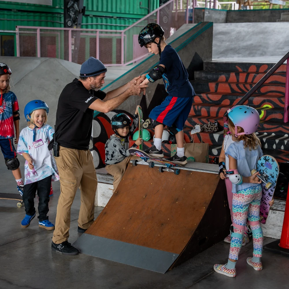 Instructor helps a young boy balance on a skateboard ramp while others wait.