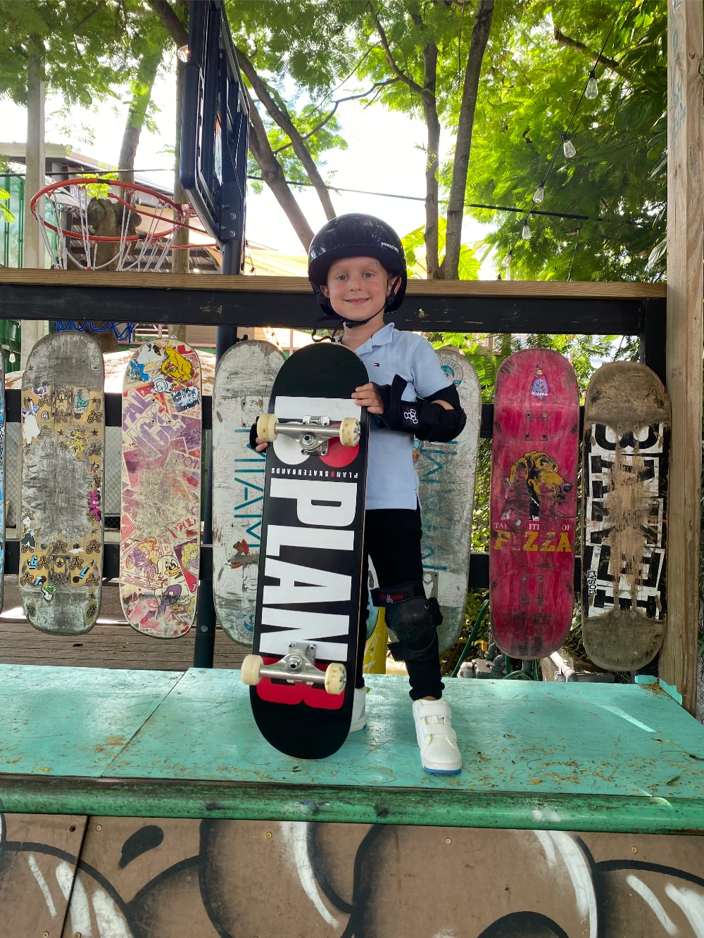 A young child in a helmet proudly holds a "PLAN B" skateboard.