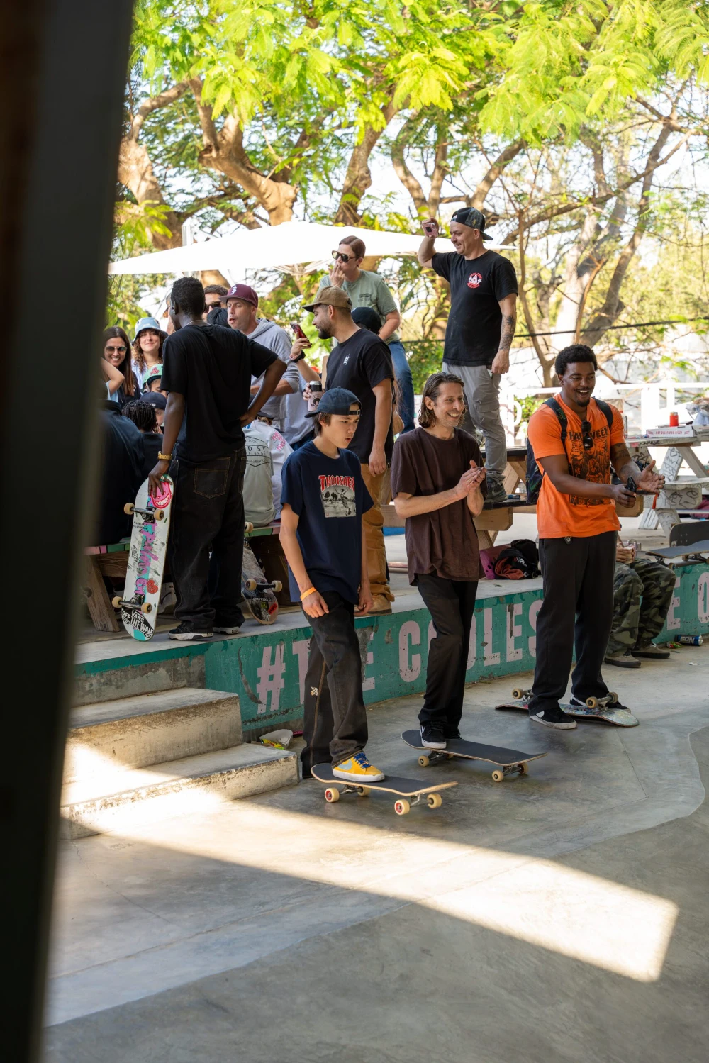A group of people watch skateboarders at an outdoor skatepark with trees.