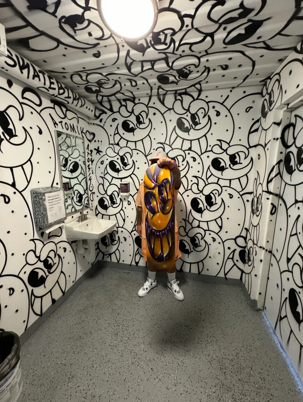 A person holds a brightly painted skateboard in a bathroom covered in graffiti.