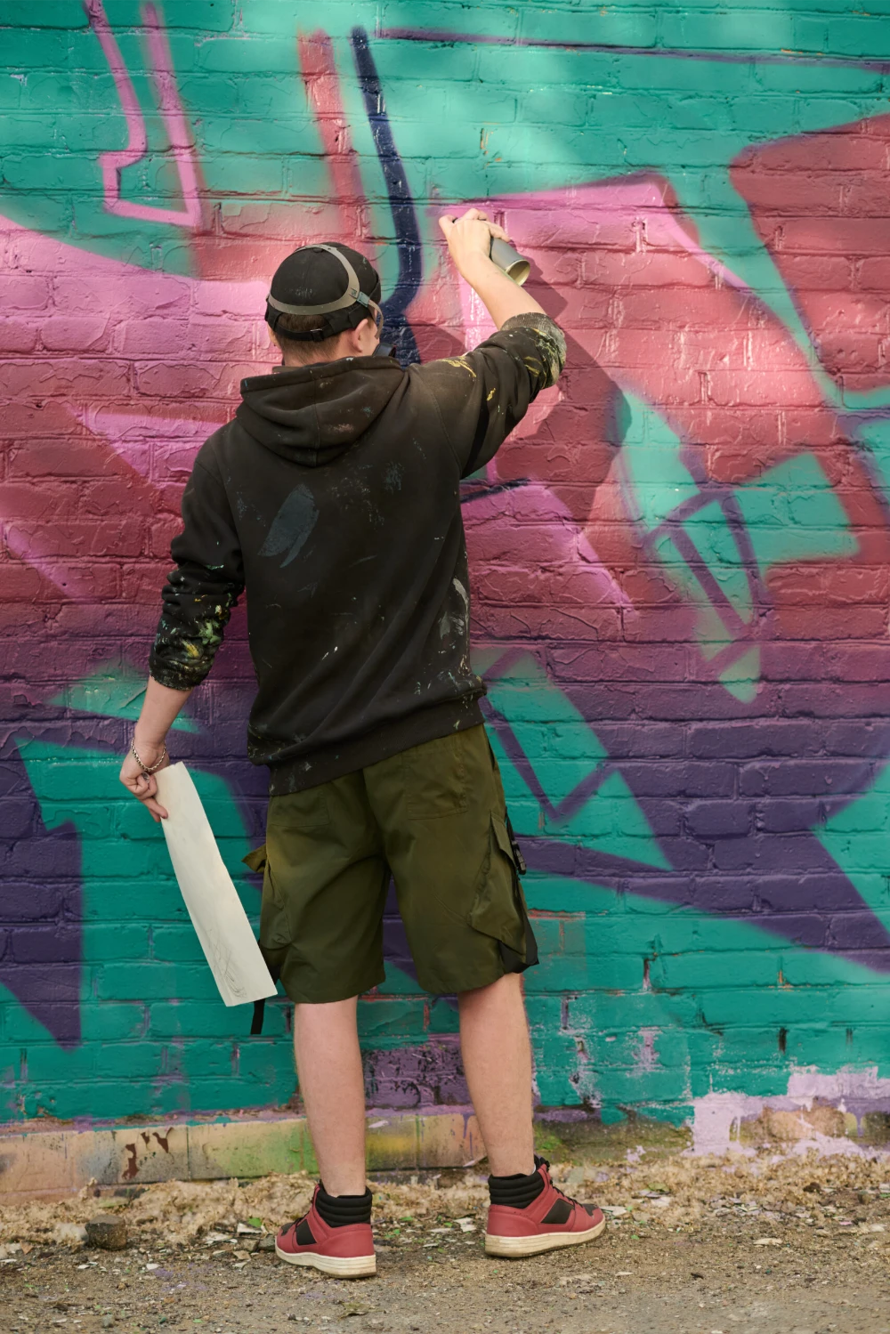 Back view of young skilled man with sketch on paper and sprayer paint creating new artwork on green brick wall at street